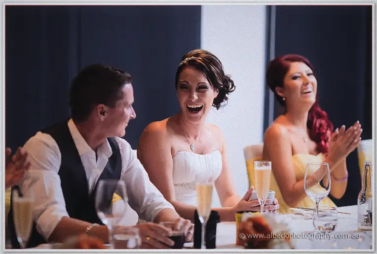 Elisha laughs at the bridal table between Shane and a red-haired bridesmaid amid champagne glasses