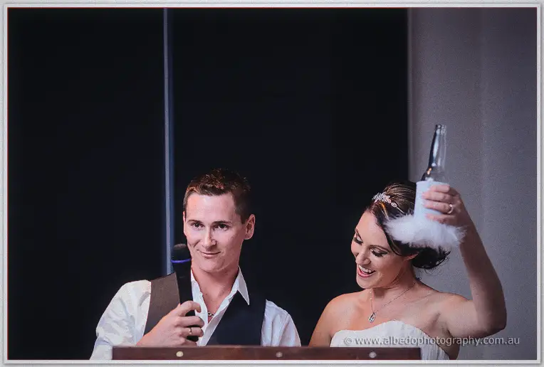 Shane grins into the mic as Elisha hoists a bottle in a fluffy white koozie during the speeches