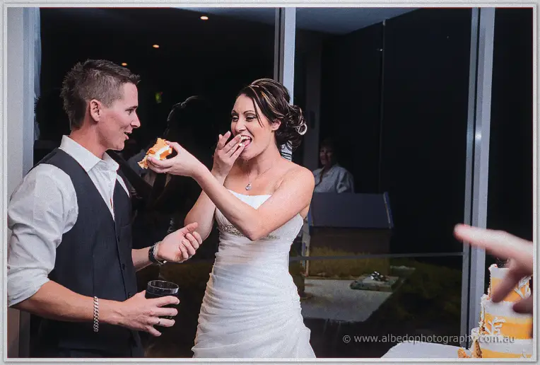Elisha licks cake from her fingers as Shane watches with his drink by the glass doors