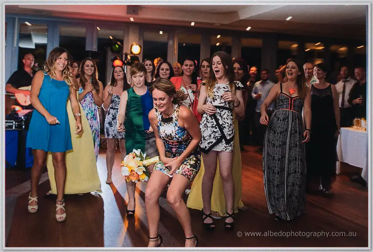 A guest in a floral dress crouches low as laughing women jostle for the bouquet at the Dawesville reception