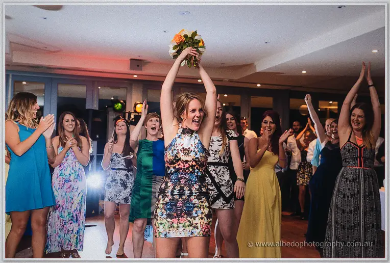 A woman in a floral halter dress lifts the caught bouquet overhead while cheering guests clap around her