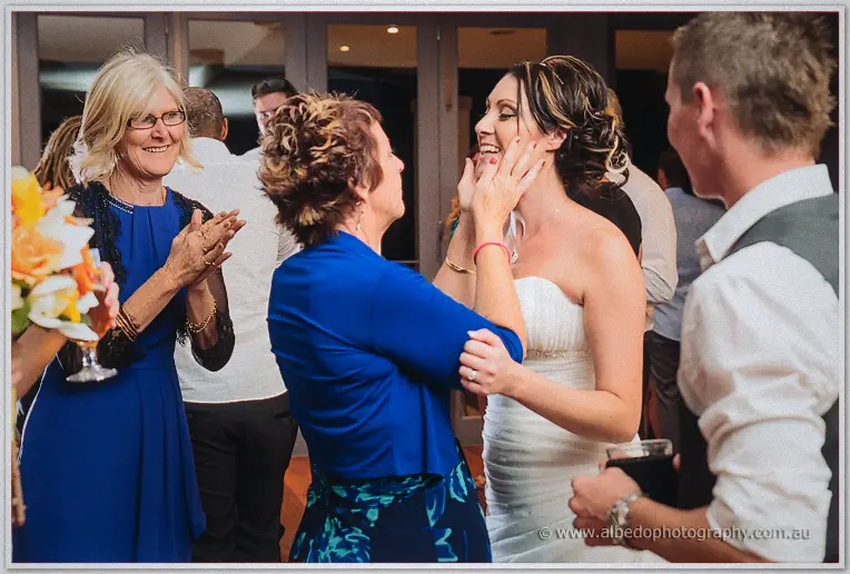 A woman in a bright blue dress holds the laughing bride's face in her hands amid clapping reception guests