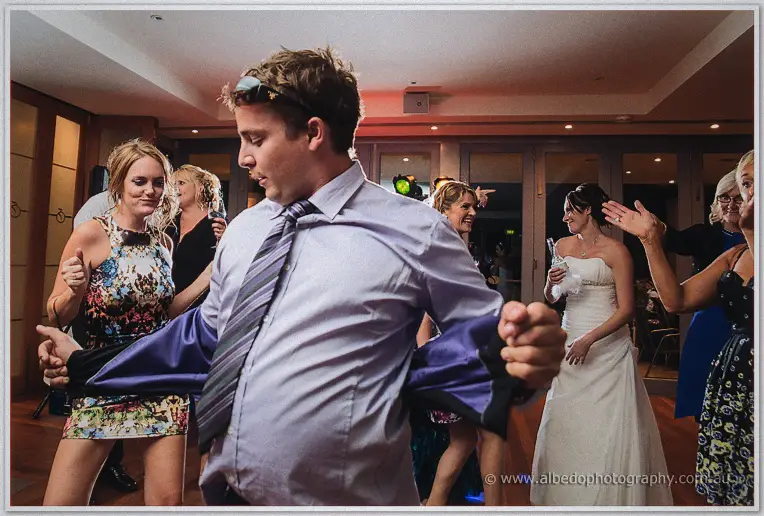 A guest in a lilac shirt and striped tie swings his jacket while dancing as the bride laughs behind him