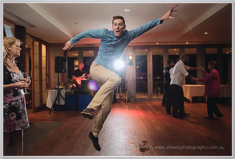 A guest in a denim shirt leaps mid-air across the glowing dance floor, arms flung wide, under party lights