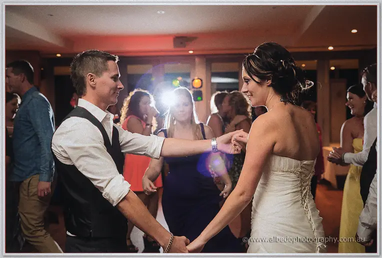 The groom in a black vest dances hand in hand with his bride, lens flare glowing between them