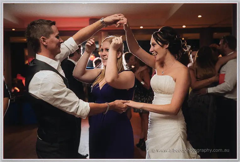 The groom twirls a laughing bridesmaid in purple beneath his raised arm as the bride beams beside them