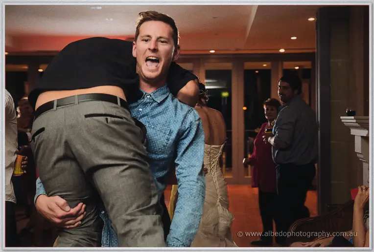 A guest in a blue shirt piggybacks a grinning mate across the floor with the bride nearby