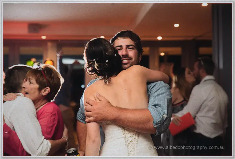 The beaming bride hugs a guest in a plaid shirt tightly at the reception while other couples embrace nearby