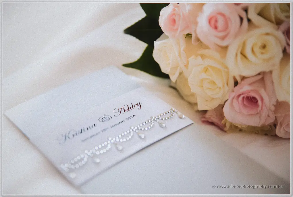 Kristina and Ashley's rhinestone-detailed invitation card beside a posy of pink and cream roses