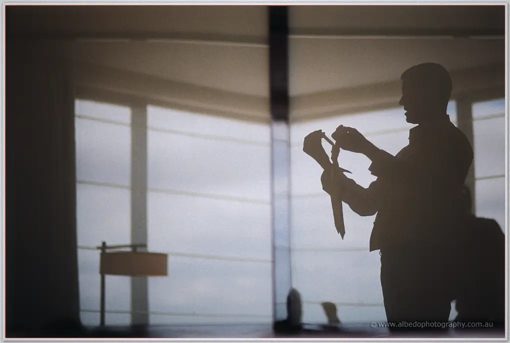 A groomsman is silhouetted at a bright hotel window as he loops his tie