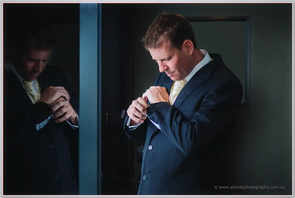 Ashley adjusts his cuff beside a dark mirror, his reflection doubling the quiet moment