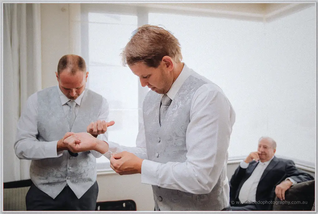 Two groomsmen in grey paisley vests fix their cuffs by the window while a guest looks on