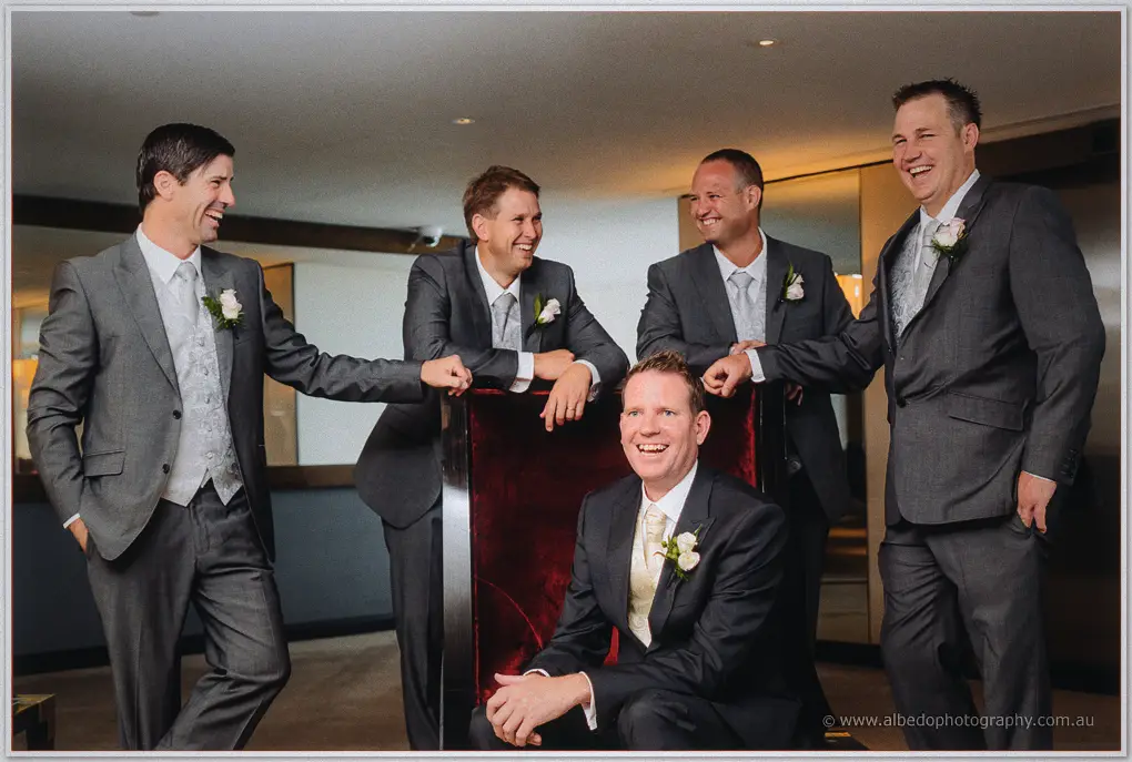 Ashley laughs from a red velvet chair, groomsmen crowding around with hands on his shoulders