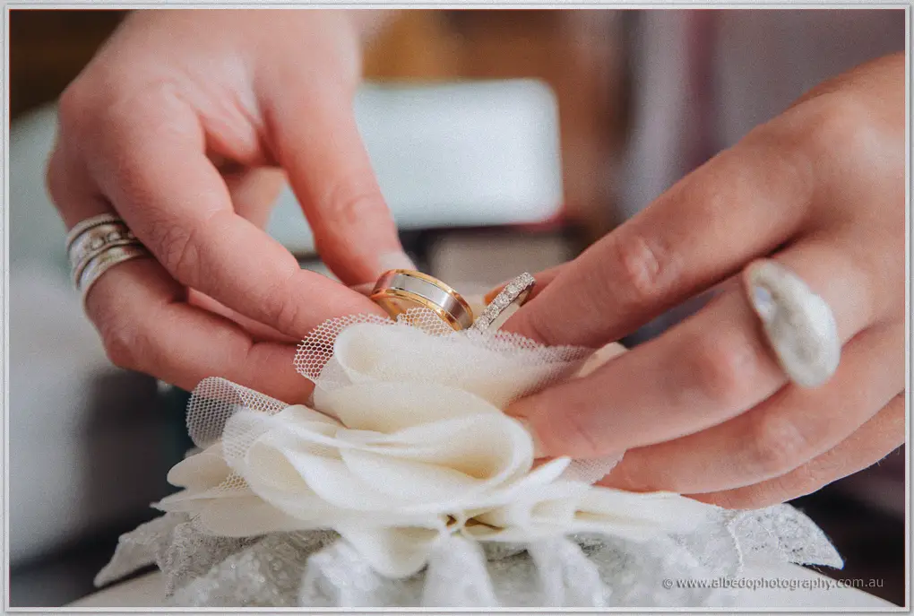 Hands nestle two wedding rings into a tulle flower on a lace pillow, macro view