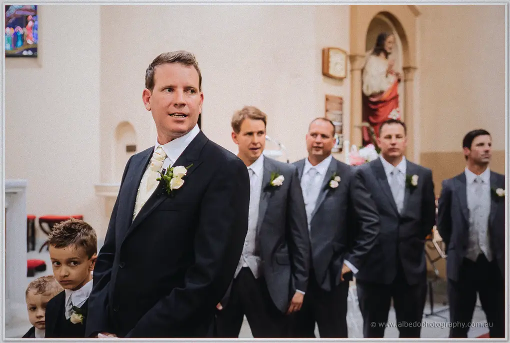 Ashley turns toward the aisle in a pale gold tie, groomsmen and page boys lined up behind
