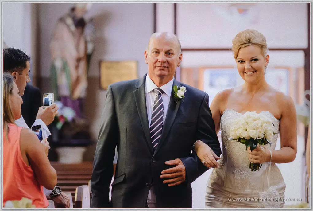 Kristina beams on her father's arm mid-aisle as a guest raises a phone to film