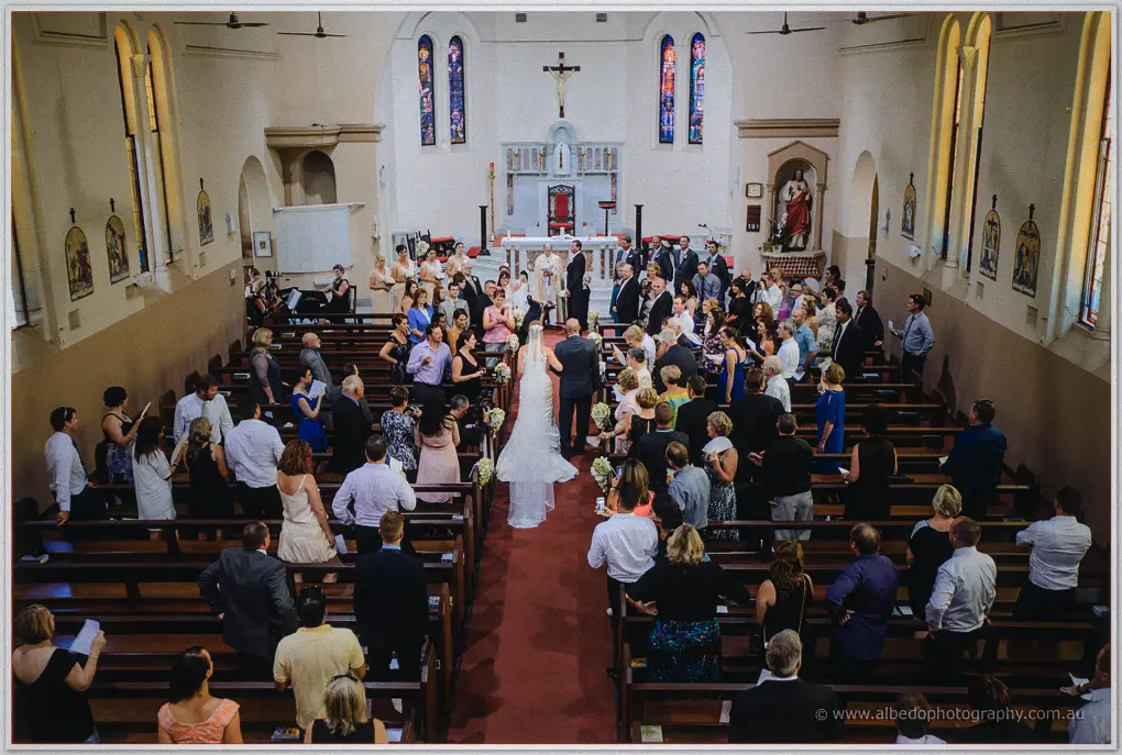 Elevated wide view of the couple before the altar as guests stand packed into the pews