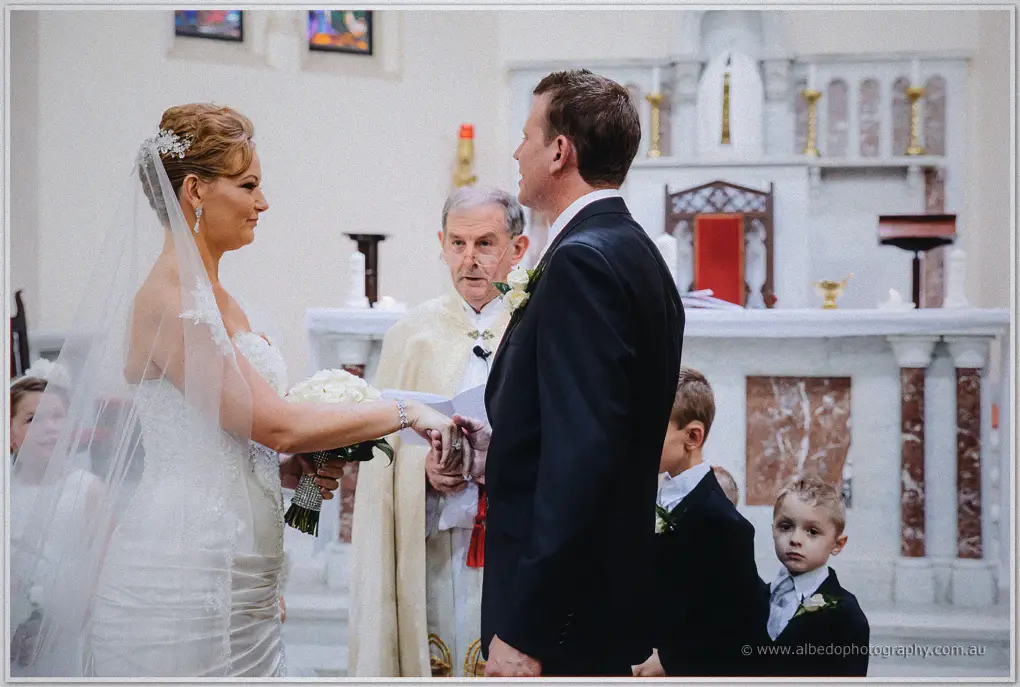 Kristina offers her hand as Ashley slides the ring on, priest and page boys beside them