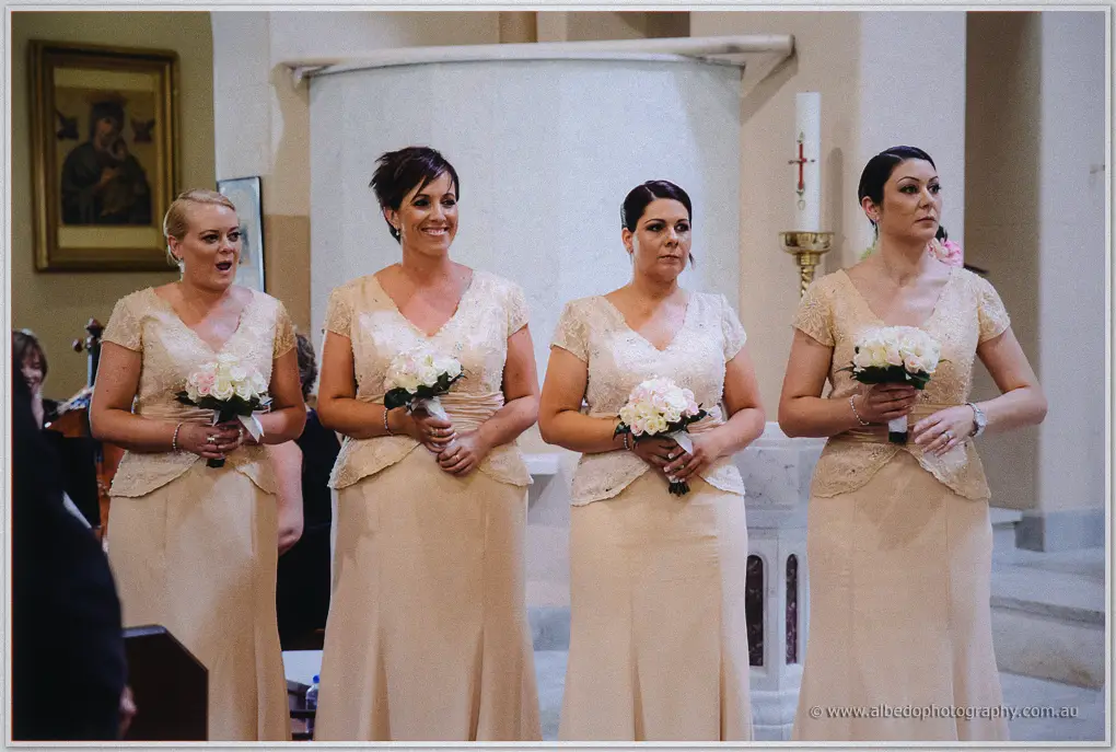 Four bridesmaids in champagne lace stand before the baptismal font with cream rose posies
