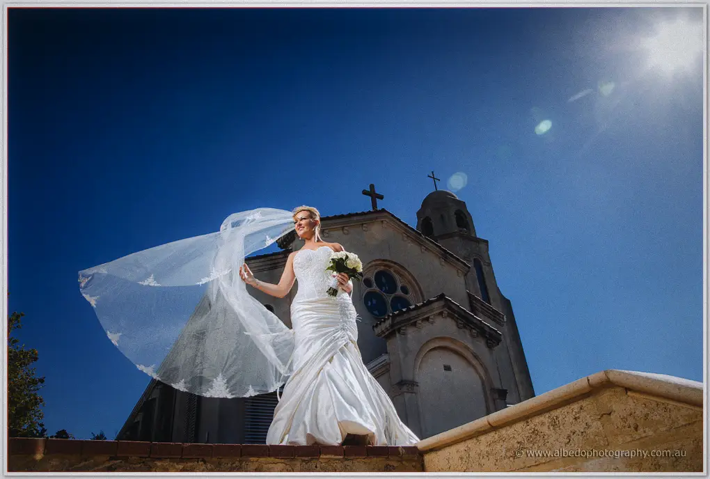 Kristina's veil billows as she holds her bouquet outside St Columba's Church under a deep blue sky