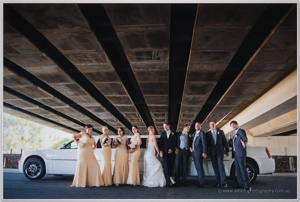 Kristina and Ashley's bridal party laughing beside a white stretch limousine beneath a concrete overpass