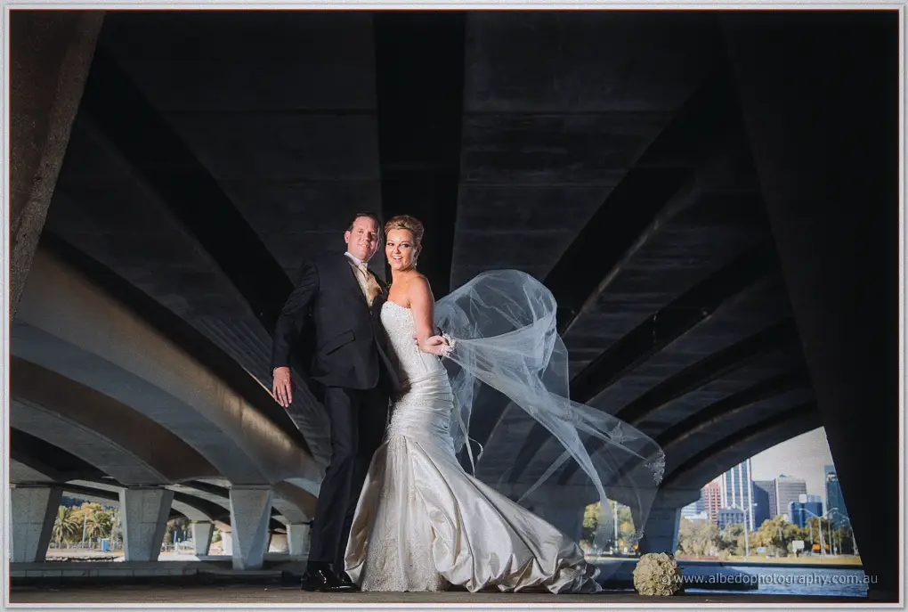 Ashley holds Kristina close beneath a dark bridge arch as her veil swirls against the Perth skyline