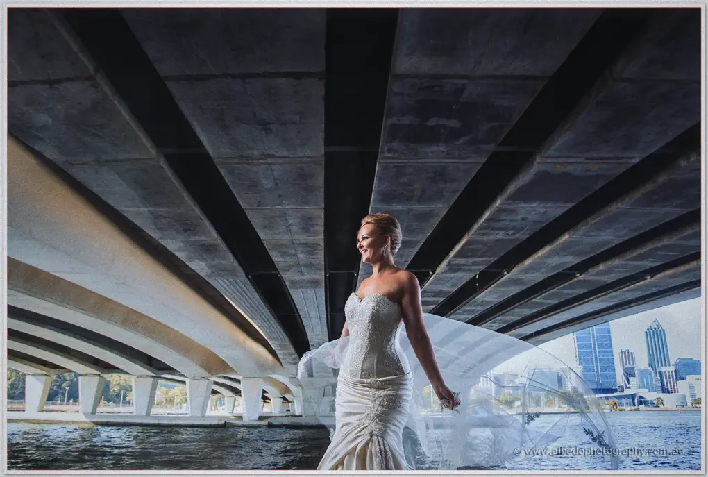 Kristina smiles beneath sweeping concrete bridge arches as her veil lifts in the breeze