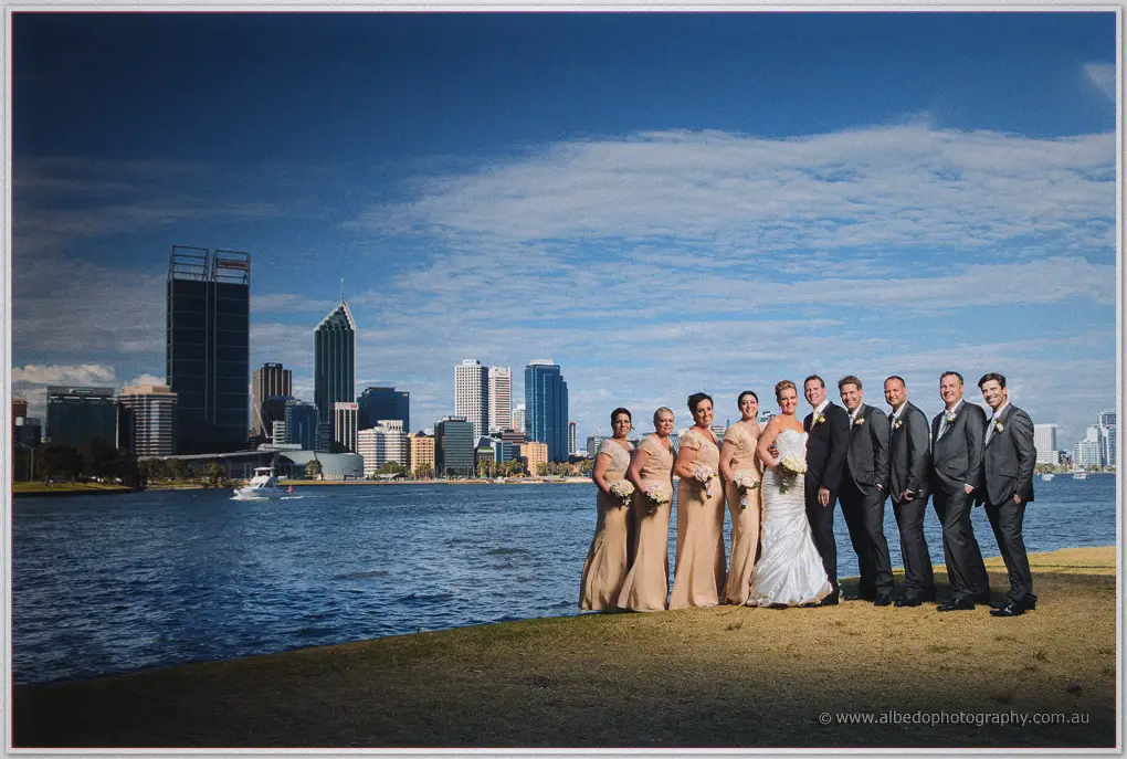Bridesmaids in champagne gowns and groomsmen in grey suits gather on the riverbank with Kristina and Ashley