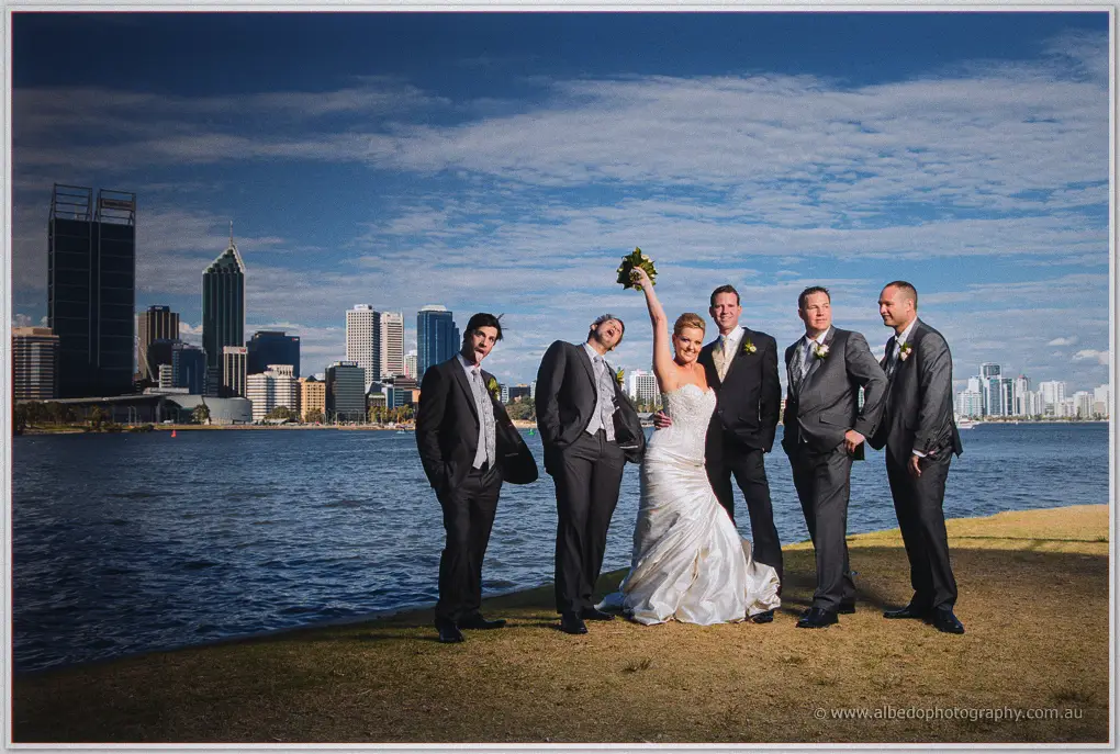 Kristina raises her bouquet triumphantly as groomsmen strike playful poses by the Swan River