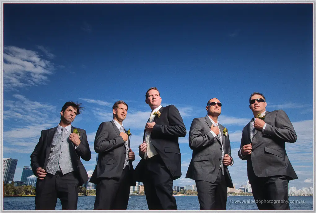 Ashley and his groomsmen adjust their jackets against a bright blue sky and the city skyline