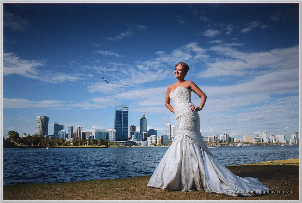 Kristina stands on the grassy riverbank, her gown trailing across the grass before the Perth skyline