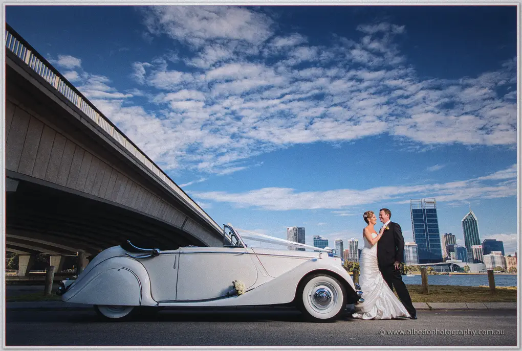 Kristina and Ashley embrace by their ribboned white convertible beneath a freeway bridge, city skyline beyond