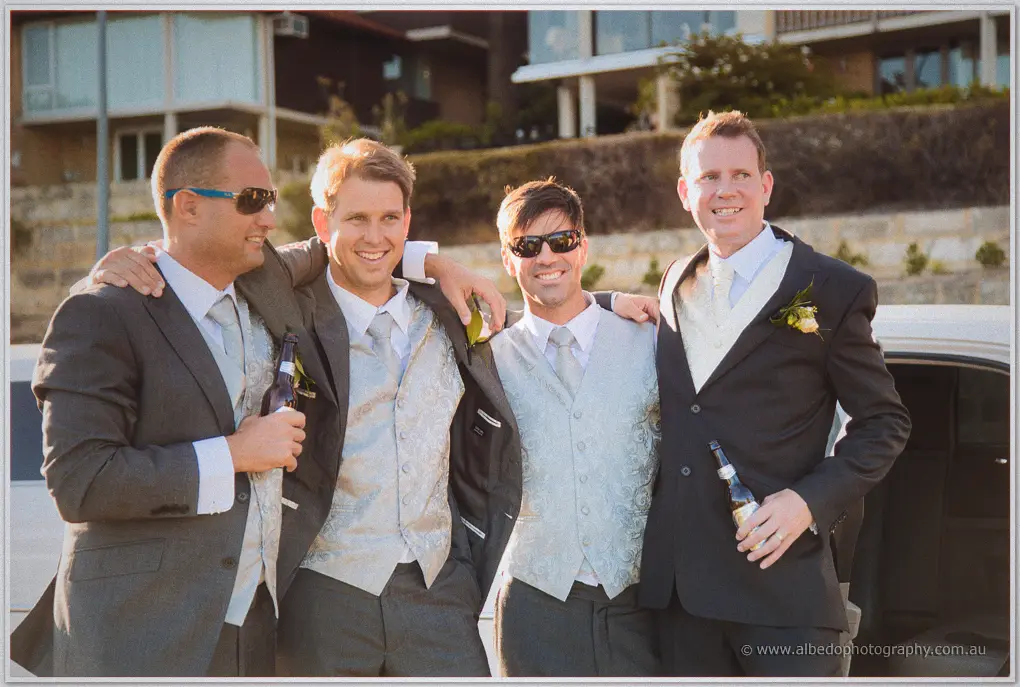 Groomsmen in silver vests share beers and laughs beside the limousine in the afternoon sun