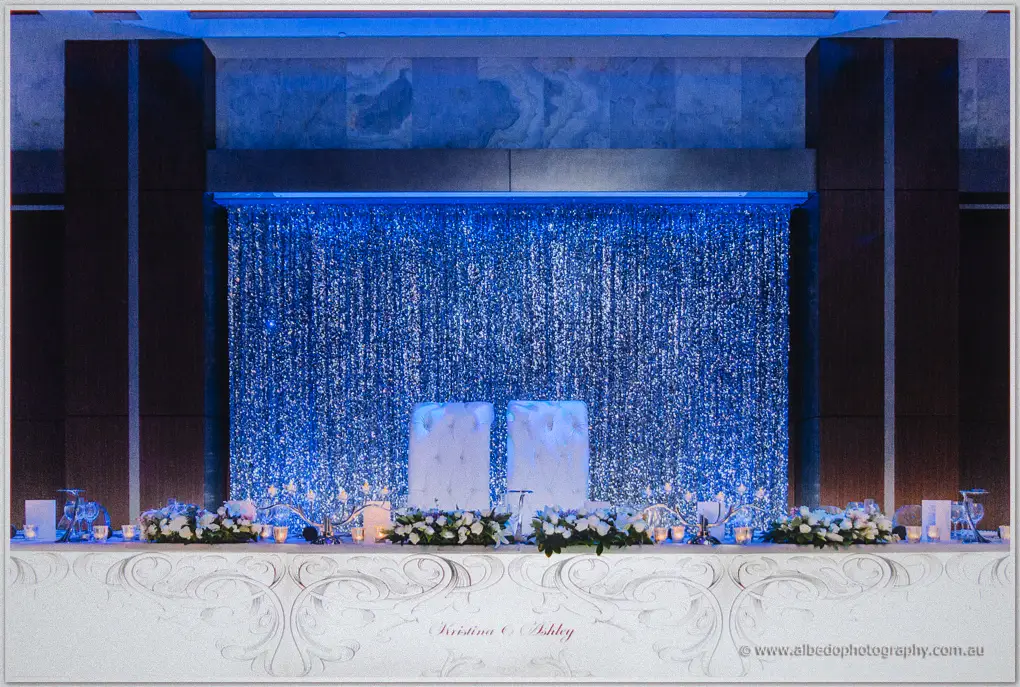 Bridal table with white thrones and rose arrangements glows before a shimmering blue curtain