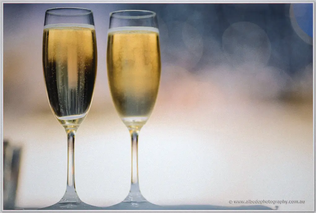 Two champagne flutes catch golden bubbles against soft bokeh by the river