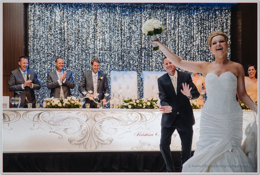Kristina raises her bouquet as Ashley dances beside the bridal table under a glittering silver curtain