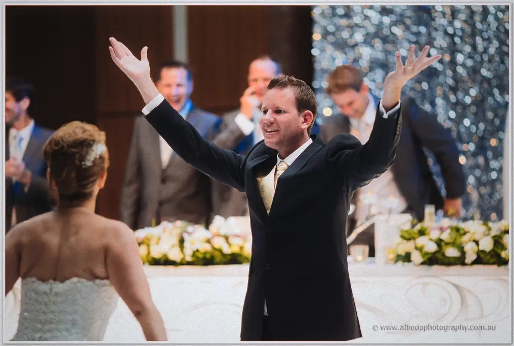 Ashley flings both arms wide in celebration as groomsmen cheer behind him and Kristina beams