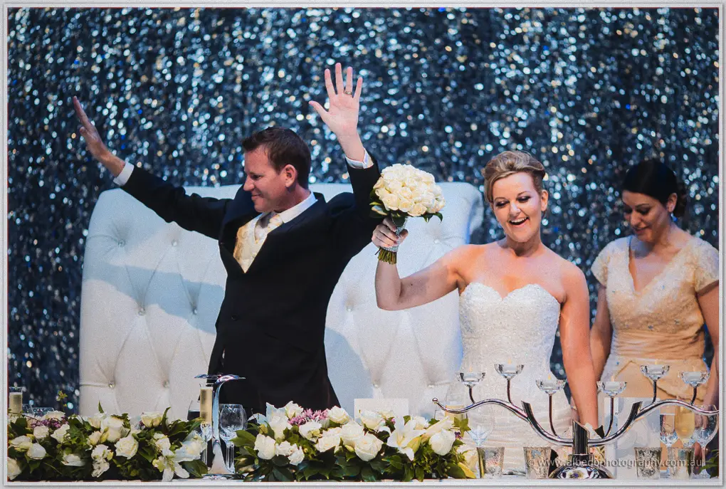Kristina raises her white bouquet as Ashley waves from the bridal table beneath silver sequins