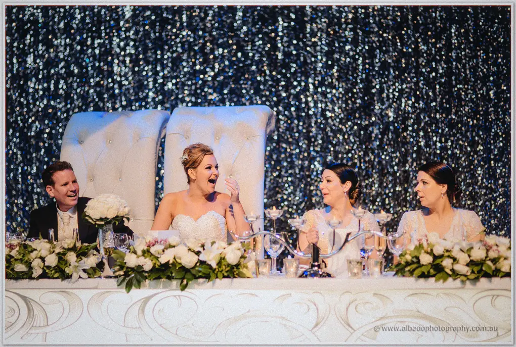Kristina gasps in delighted laughter at the bridal table flanked by her bridesmaids