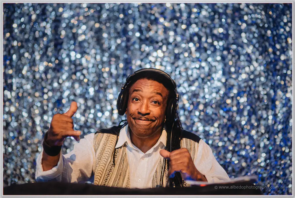Smiling DJ with dreadlocks and headphones spinning tracks in front of a sparkling silver backdrop.