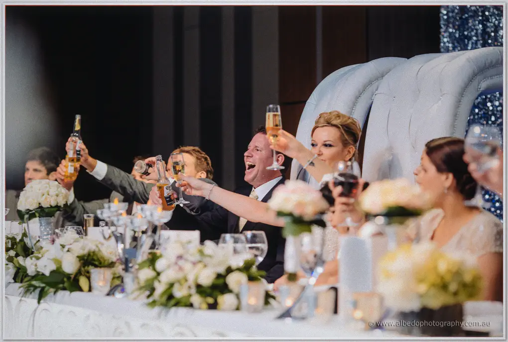 Kristina and Ashley toast with champagne and beer amid white roses on the bridal table