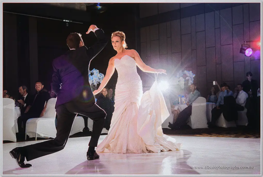 Ashley crouches playfully before Kristina as laughing guests watch from tables at their Crown reception.