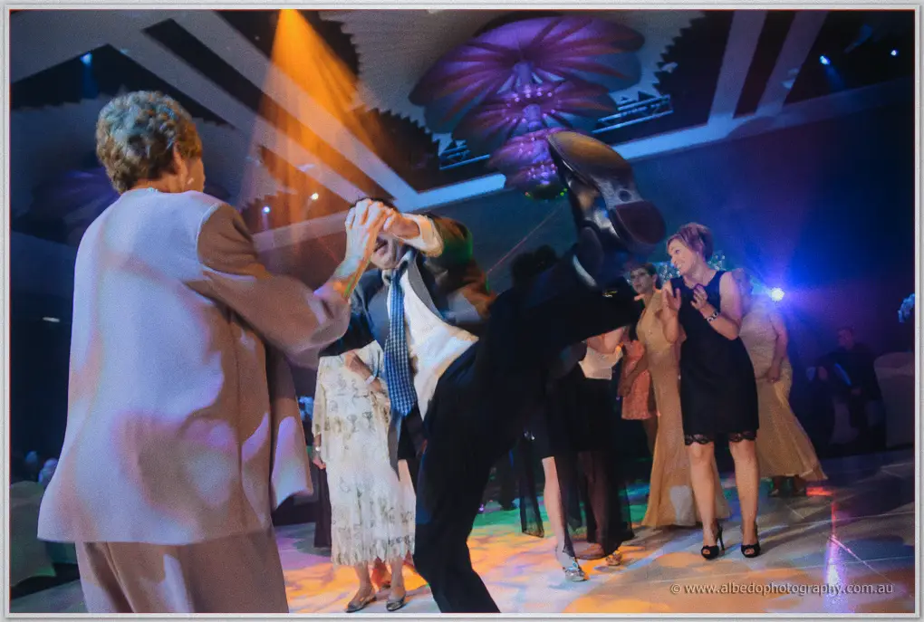 A guest kicks his leg high while dancing with an older woman under warm spotlights at the Astral Ballroom.
