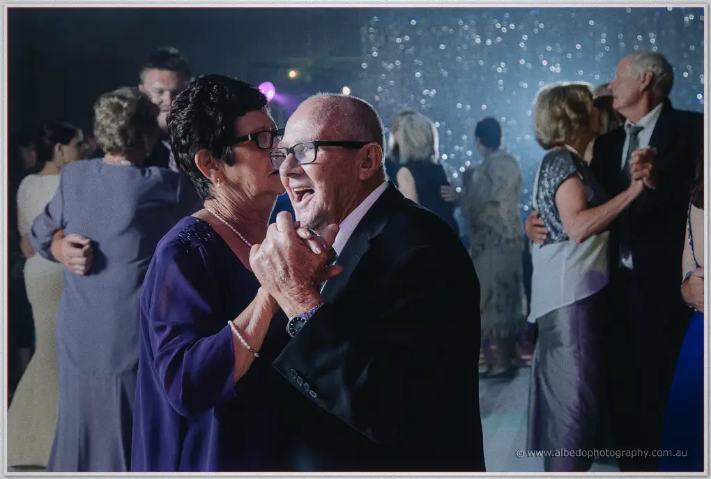 An elderly guest in purple kisses her laughing partner's cheek during a slow dance under the sparkle backdrop.