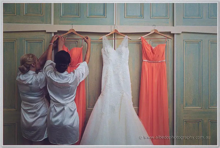 Bridesmaids in silk robes hang coral dresses beside Katelyn's lace gown on vintage teal doors