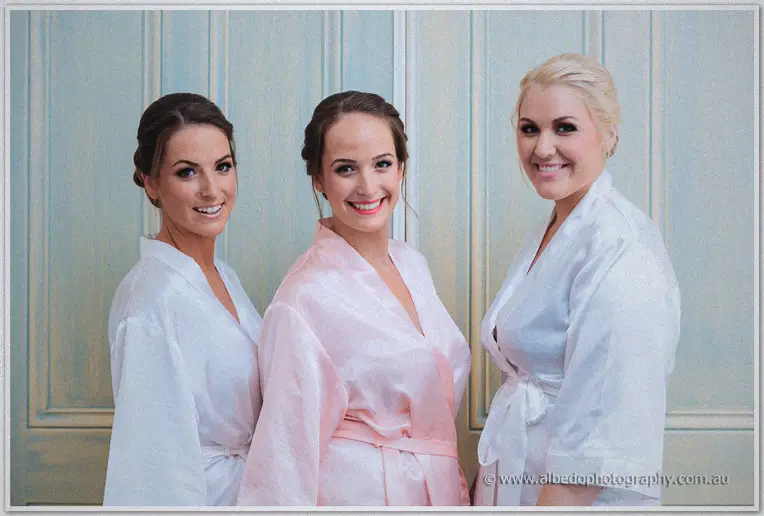 Katelyn flanked by two bridesmaids in silk robes, all smiling against pale blue panelling