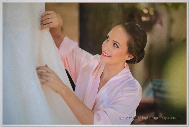 Katelyn adjusts the neckline of her lace gown in a pink robe before a garden window