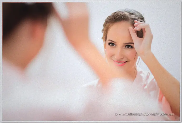 Katelyn tucks a strand of hair into her updo while meeting her reflection, soft morning light