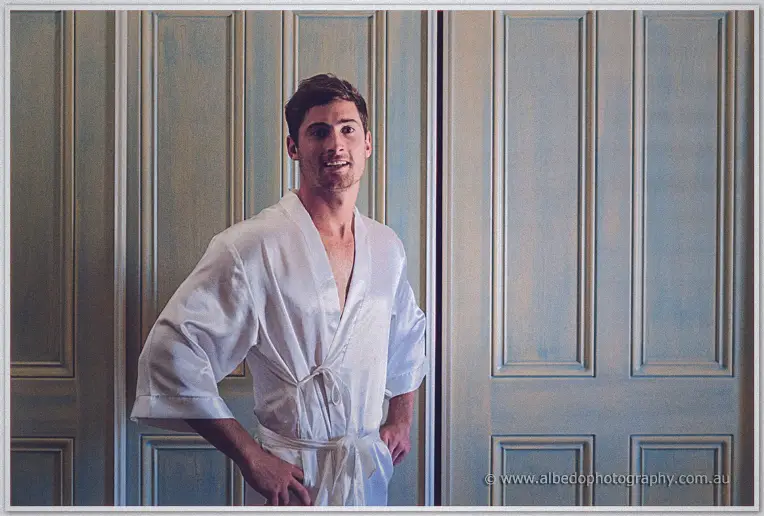 Matt plays superhero in a white satin robe, hands on hips, grinning before the wardrobe