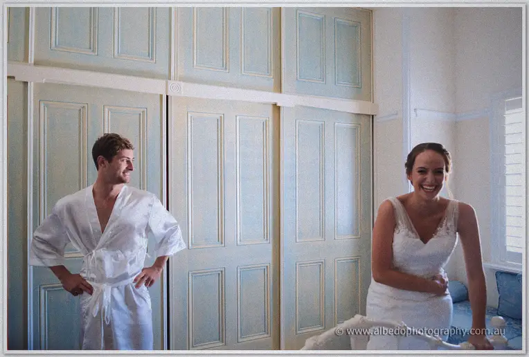 Matt in his robe and Katelyn in her gown laugh together in the bright bridal room